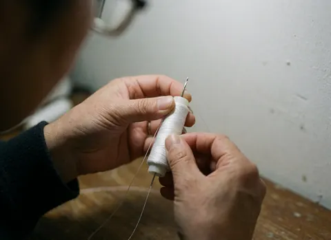 Close-up of white thread being stitched with a metal needle on a wooden surface against a blurred white wall.
