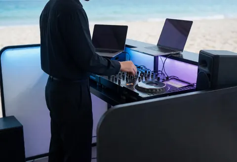 Professional DJ adjusting a mixer in a beach cabana with blue and purple lighting, laptops, and speakers against a blurred beach background