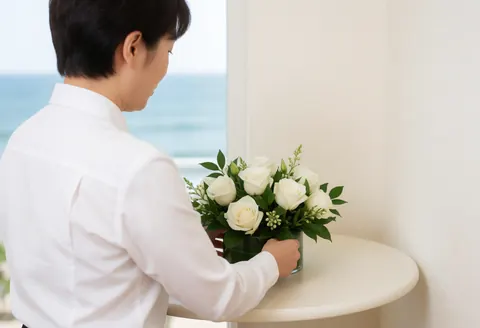 Staff member adjusting a white rose floral arrangement on a round table near a window with a sea view in a bright beach club setting