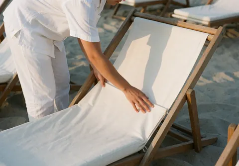 Worker in white uniform adjusts a wooden hammock cushion on light sand under warm sunlight