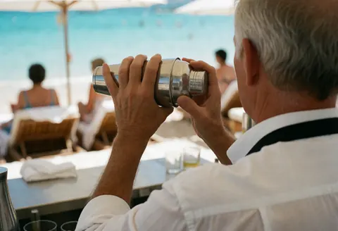 Bartender shaking cocktail shaker at beach, back view, hands and tool visible, blurred background with water and chairs