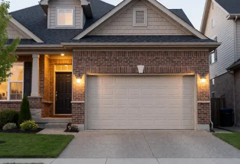Residential house with closed beige garage door, brick facade, warm entrance lighting, and well-maintained garden.