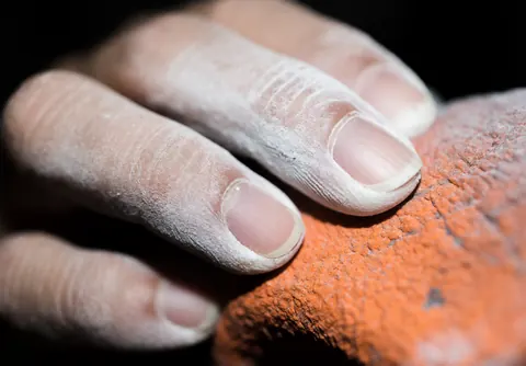 Close-up of an adult hand covered in white chalk gripping a rough orange rock with selective focus and direct lighting
