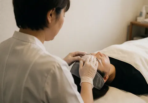 Therapist wearing gloves applies white cream to client's forehead while client rests with gray headband in calm, professional spa setting