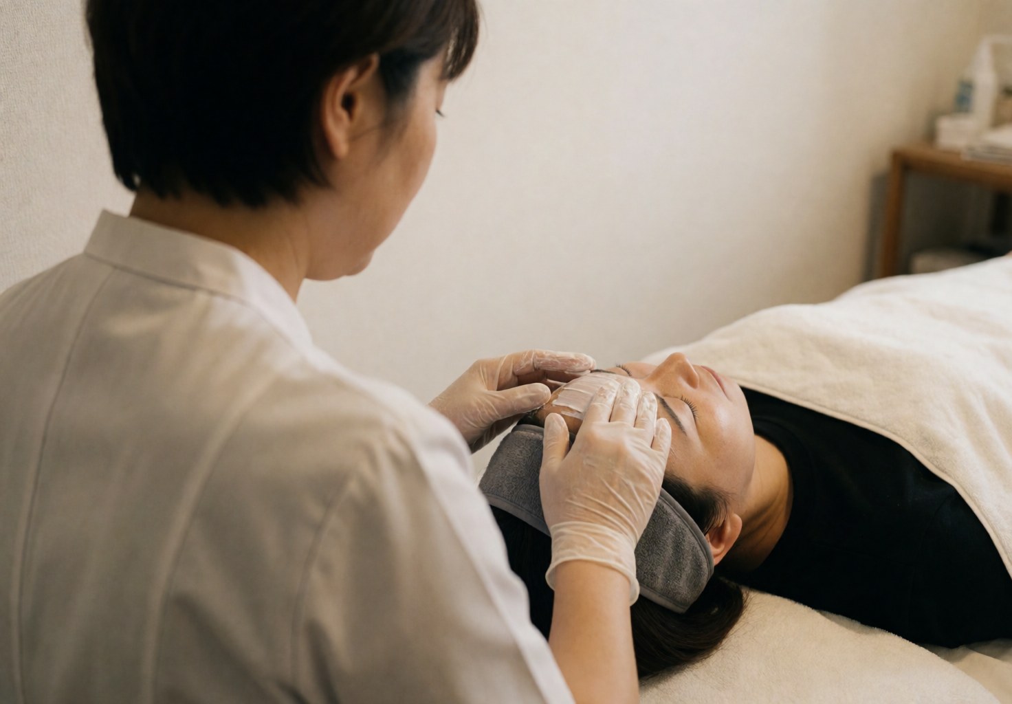 Therapist wearing gloves applies white cream to client's forehead while client rests with gray headband in calm, professional spa setting