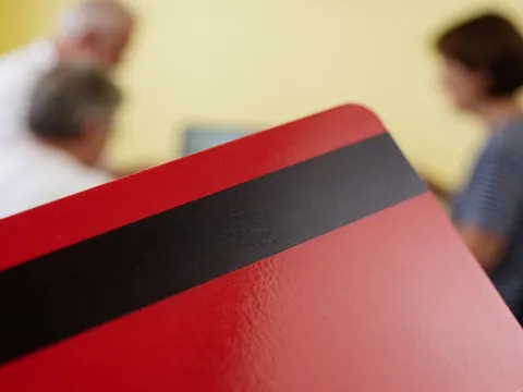 Red plastic card featuring a black magnetic stripe, displayed against a blurred yellow wall background with indistinct figures nearby