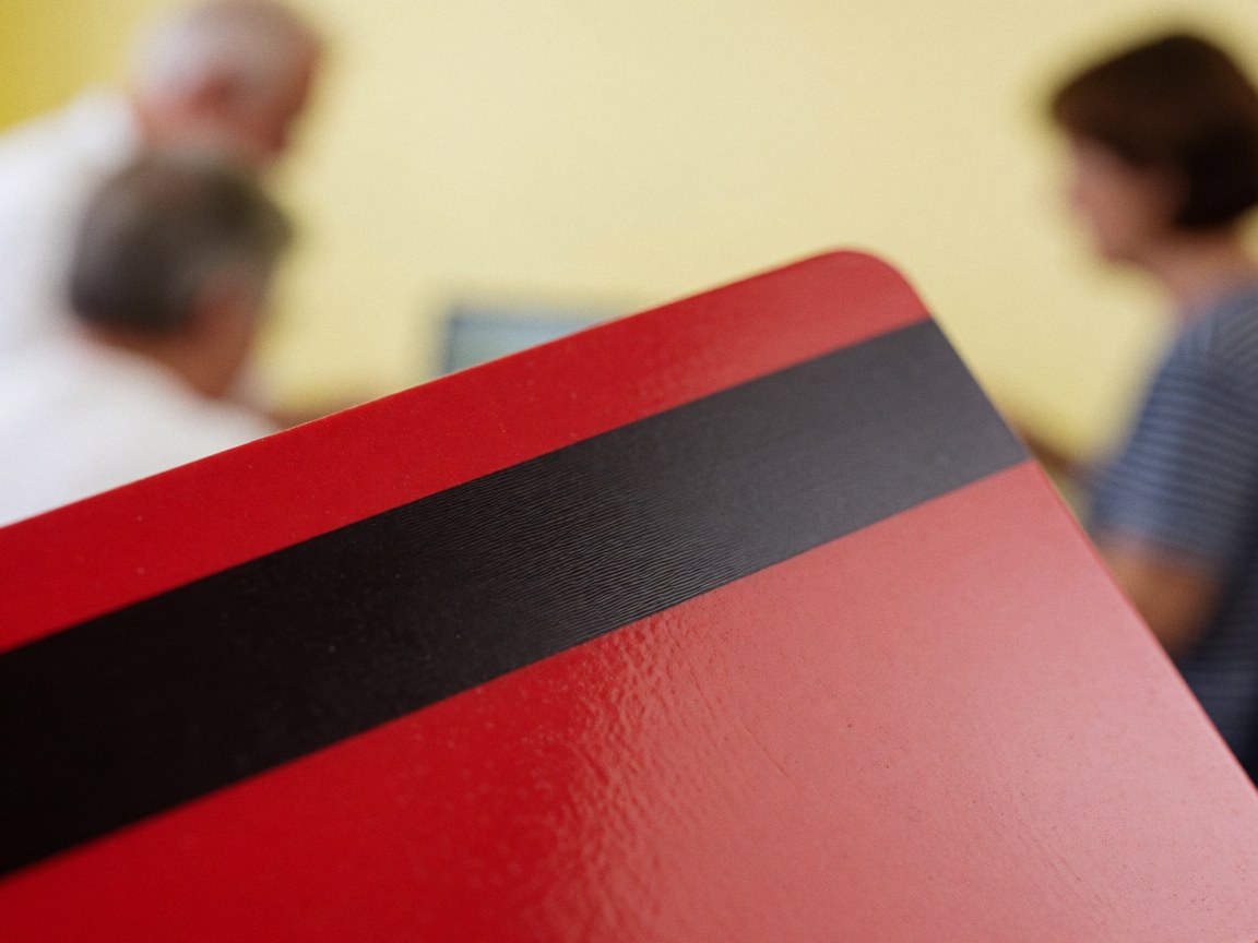 Red plastic card featuring a black magnetic stripe, displayed against a blurred yellow wall background with indistinct figures nearby