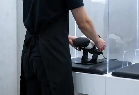 Staff member placing a VR headset on a charging station behind a clear plastic barrier in a modern entertainment center.