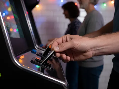 Close-up of a hand inserting a card into a black arcade machine slot with blurred party lights and white tiled walls in the background.