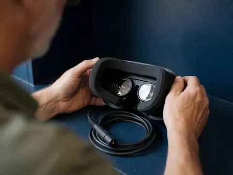 Close-up of an adult's hands adjusting a wired virtual reality headset on a dark blue surface, showing the lenses and foam padding.