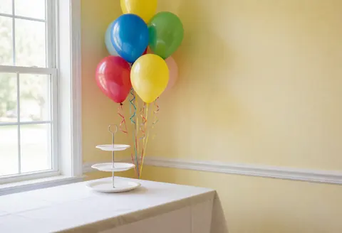 Colorful balloons tied with ribbons sit near an empty cake stand on a table with a white tablecloth in a yellow room with natural window light.