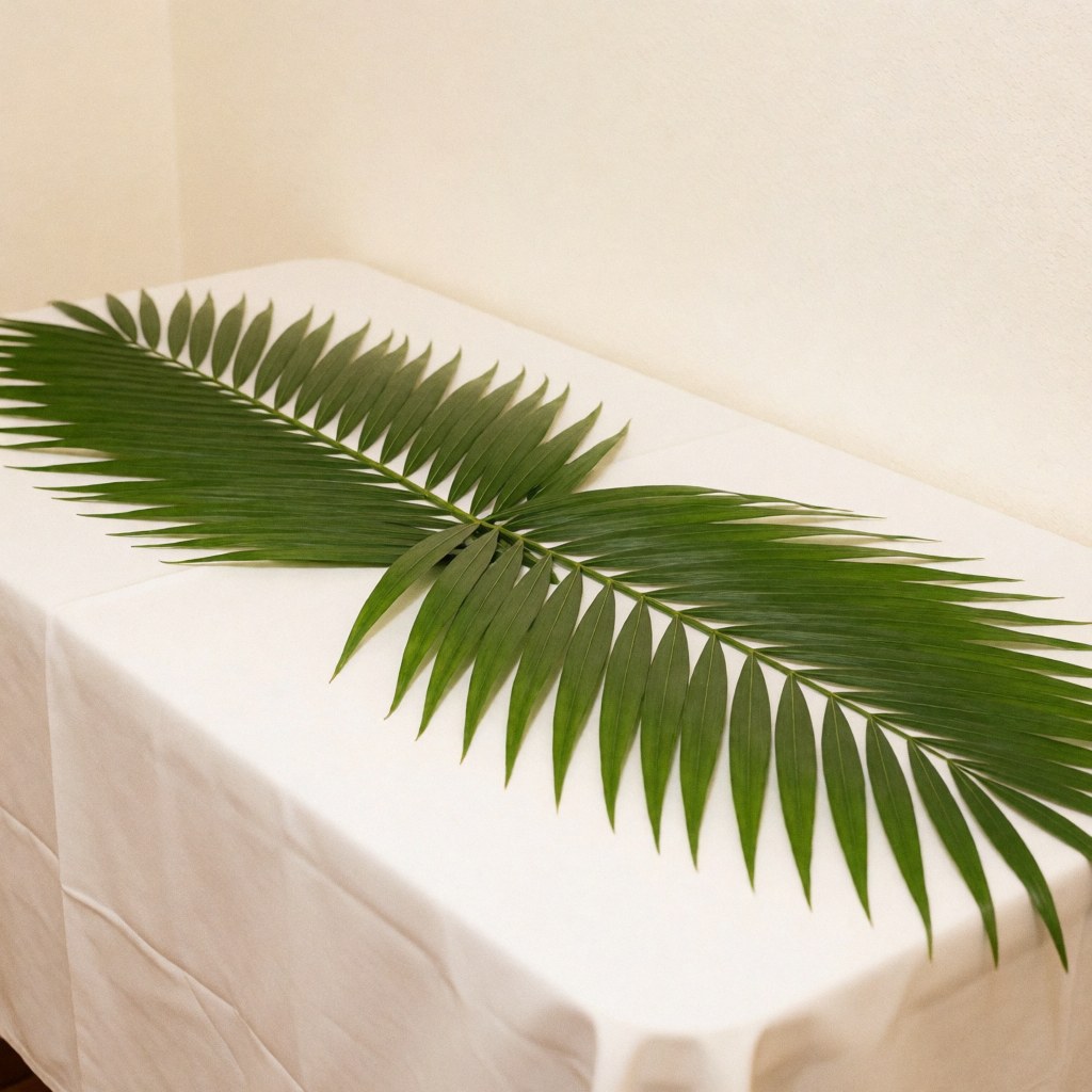A long green palm leaf placed on a white tablecloth, suitable for elegant banquet and event decoration with a natural ambiance.