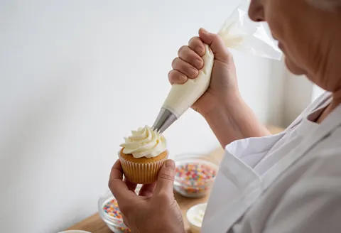 Close-up of senior hands applying white frosting to a cupcake in a professional cooking class setting
