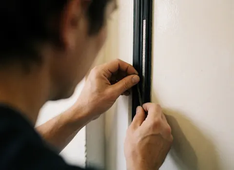 Close-up of technician hands using a tool to adjust the magnetic gasket on a refrigerator door against a warm white wall.