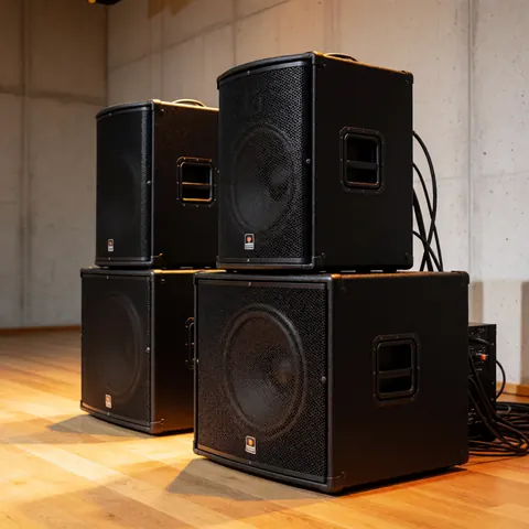Black professional party speakers with grilles placed on a wooden floor against concrete walls with warm lighting for large events