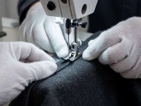 Close-up of gloved hands using an industrial sewing machine to stitch dark gray textile, highlighting metallic details and precise craftsmanship.