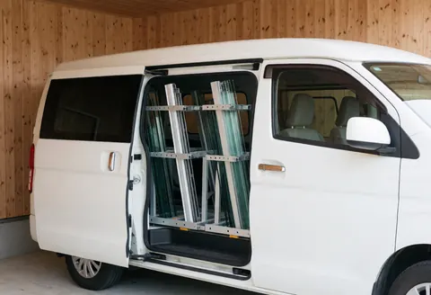 White service van parked in a garage with open door showing racks of glass panels and aluminum profiles