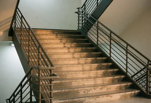 Close-up of dirty metal commercial stairs with railings, white walls, and warm lighting in an office building interior.