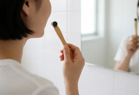 A person stands in a white bathroom using a wooden toothbrush for oral hygiene while looking into a mirror with natural daylight.