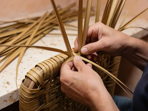 Close-up of hands weaving palm leaves into a traditional wooden chair frame in a workshop setting