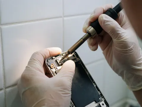 Close-up of gloved hand using a soldering iron to repair a 3.5mm jack on a smartphone against a white tiled wall with visible circuit details