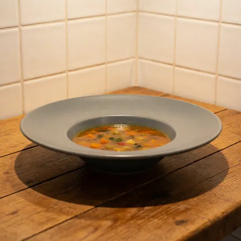 Bowl of vegetable soup in a matte gray ceramic dish placed on a rustic wooden table with white wall tiles in the background and warm cozy lighting.