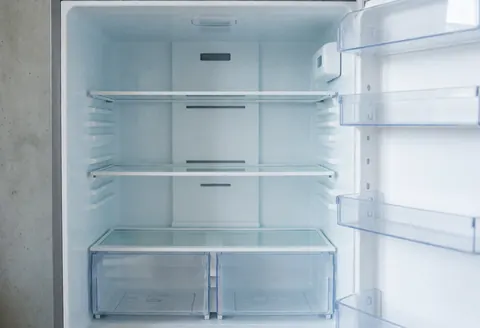 Clean interior of an empty refrigerator showing transparent shelves and drawers against concrete walls with natural light.