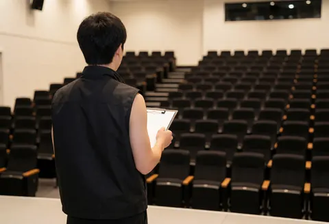 Technician in black vest checking empty auditorium with board, black chairs, white walls, lit stage.