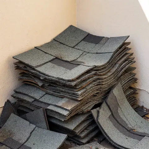 Old cracked and worn asphalt shingles stacked in a beige wall corner showing granular texture and uneven layers