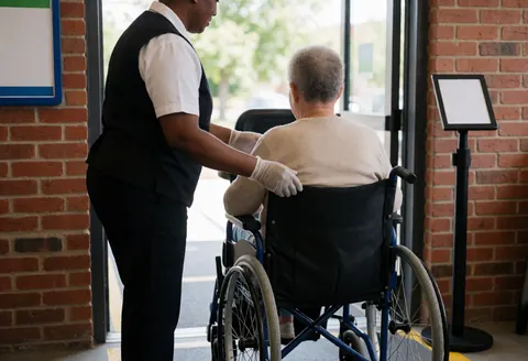 Theme park staff member wearing gloves helps an older customer in a wheelchair near a brick wall entrance.