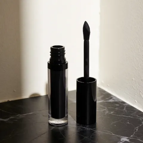 Black cosmetic liquid in a tube with an applicator resting on a dark marble surface with dramatic side lighting for professional makeup product display