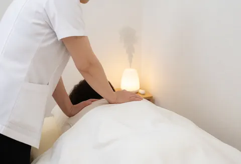 Therapist in white uniform massaging client's back covered with white sheet, using aroma diffuser emitting vapor in warm lit room