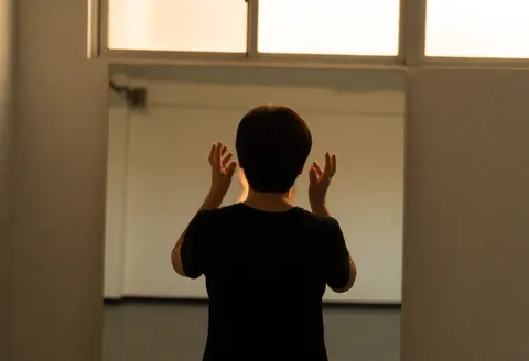 Back view of person rehearsing theater gestures in front of mirror in bright rehearsal room with warm light from high window