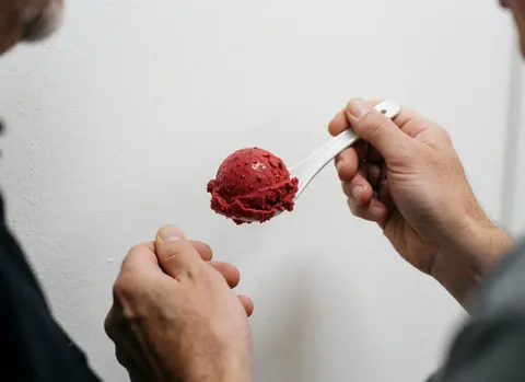 Close-up of raspberry ice cream with visible seeds served in a small white ceramic spoon held by hands against a plain white background