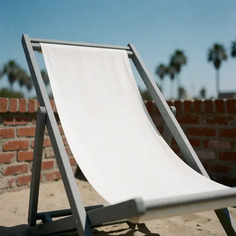 White beach chair with gray frame on sand, red brick wall and blurred palm trees under blue sky