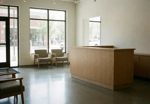 Clean minimalist hostel lobby featuring a light wood reception counter, waiting chairs, and large windows with natural light