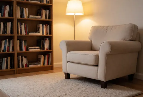 A comfortable beige armchair sits on a white rug next to a wooden bookshelf filled with books, illuminated by a floor lamp in a warm, inviting space.