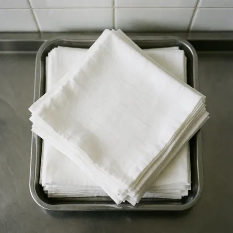 Stacked sterile white gauze pads on a stainless steel tray atop a polished metal surface with white tiled walls in the background