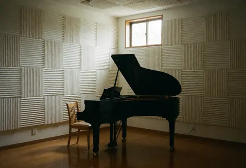 Empty music practice room featuring a black grand piano, wooden chair, and acoustic wall panels illuminated by natural window light.