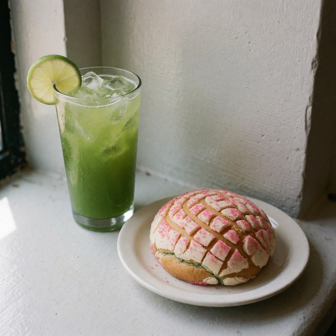 Green fresh water with lime slices and ice in a tall glass, next to a pink sugar shell on a white plate by a window with natural light.