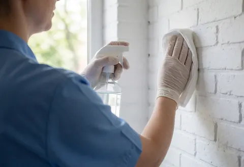 Therapist wearing white gloves cleans a white brick wall with a cloth and disinfectant spray near a window.
