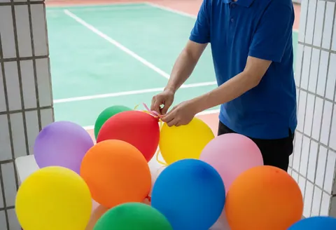 A professional in a blue shirt arranges colorful balloons on an outdoor sports court for an event.