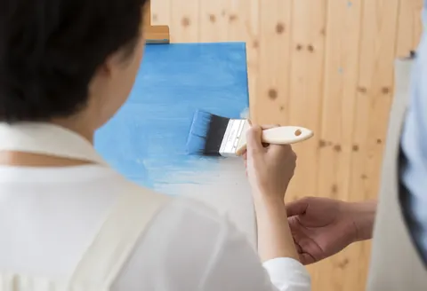 An adult student paints on a blue canvas while an instructor's hand guides their arm in a workshop with light wood walls