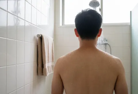 Shirtless man standing under shower in bathroom with white tile walls, beige towel hanging on left, natural light from window