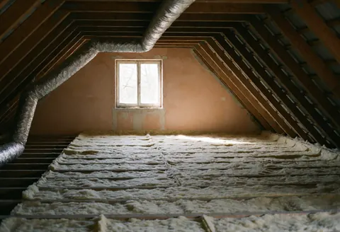 Construction ceiling space with fiberglass thermal insulation between beams, metal ducts, and pink drywall walls, illuminated by a window.