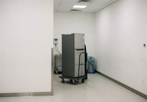 Two-door refrigerator on a hand truck inside a clean repair shop with white walls and organized tools for appliance service