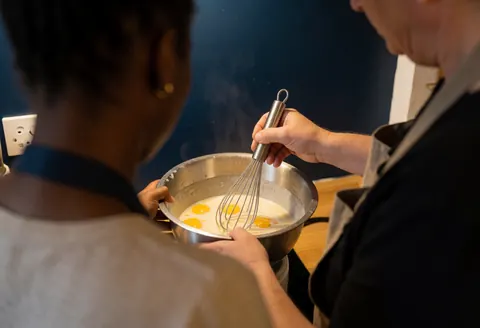 Instructor guiding a student to mix eggs in a kitchen with dark blue walls and warm lighting