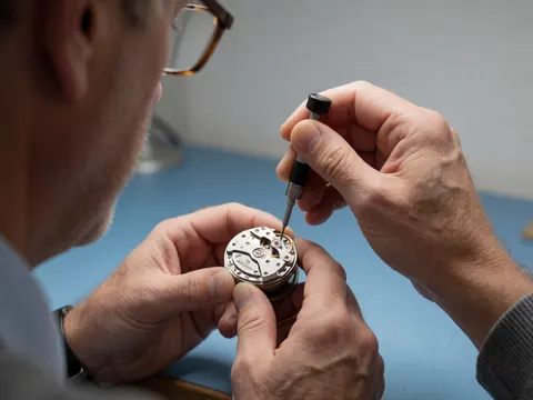 Close-up of a hand using a specialized tool to adjust the balance of a watch movement on a blue surface.