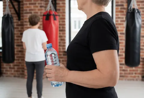 Woman holding a water bottle in a gym with boxing bags and brick wall background