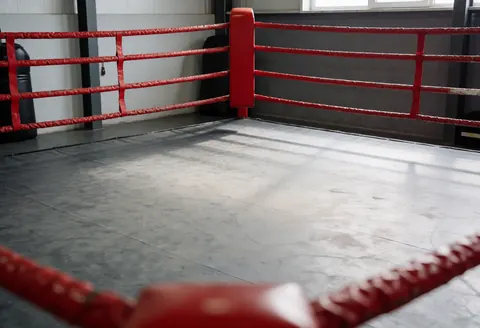 Empty boxing ring with red ropes, concrete floor, and gray walls suitable for training